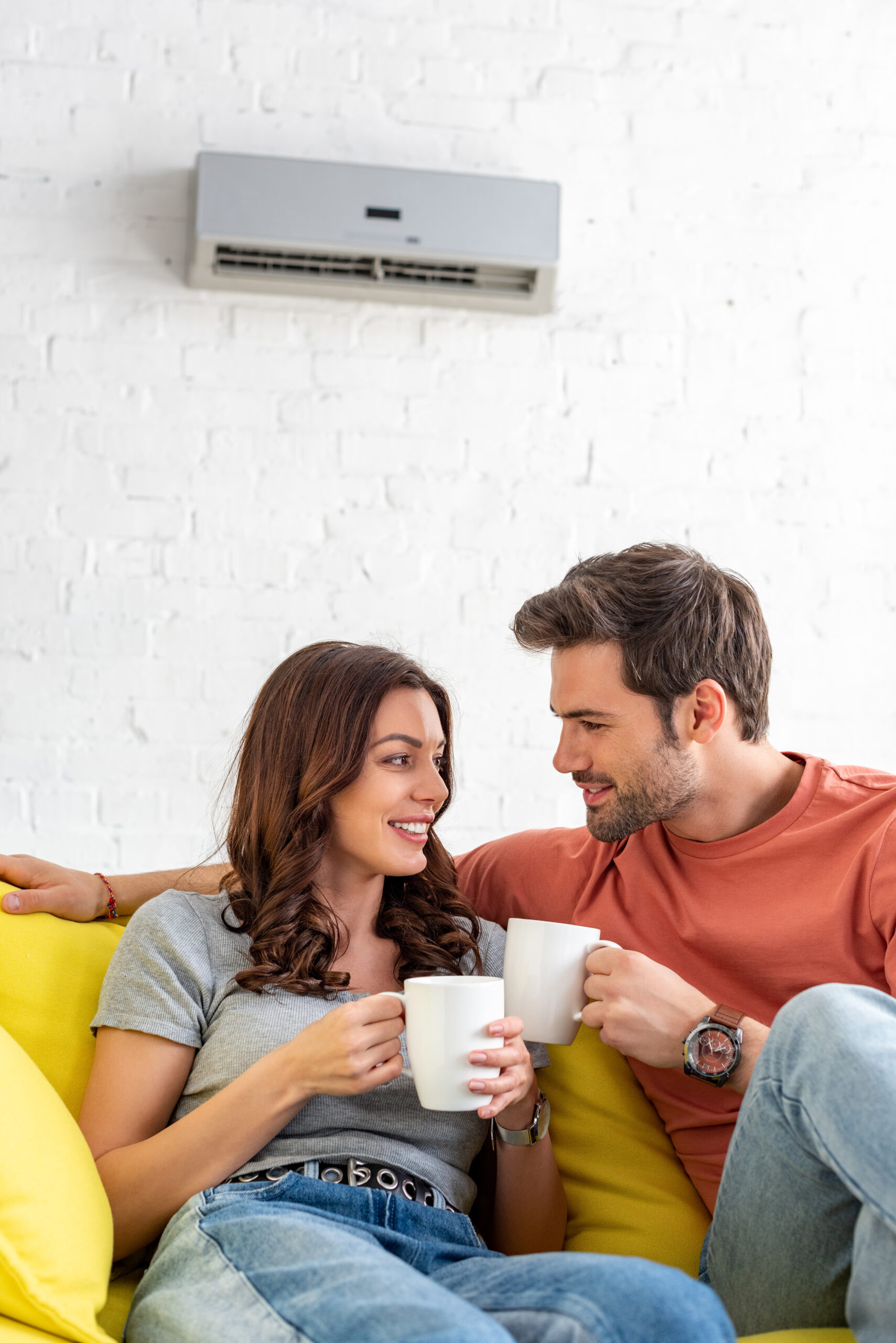 happy smiling couple holding cups while sitting under air conditioner at home
